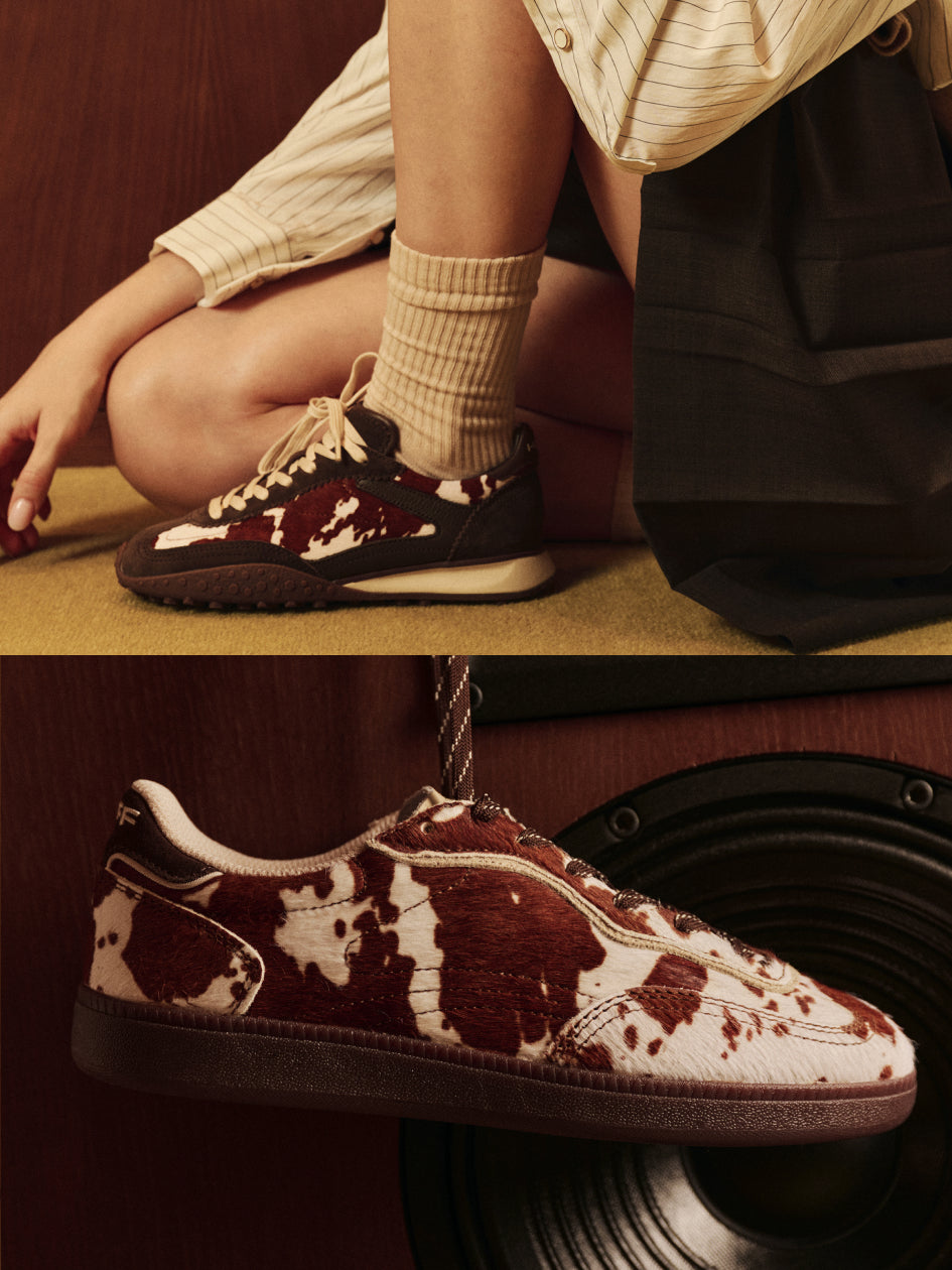 A pair of low-top sneakers with a brown and off-white cow-print pattern, shown worn on a person's foot in an indoor setting and also hanging against a dark wooden background.